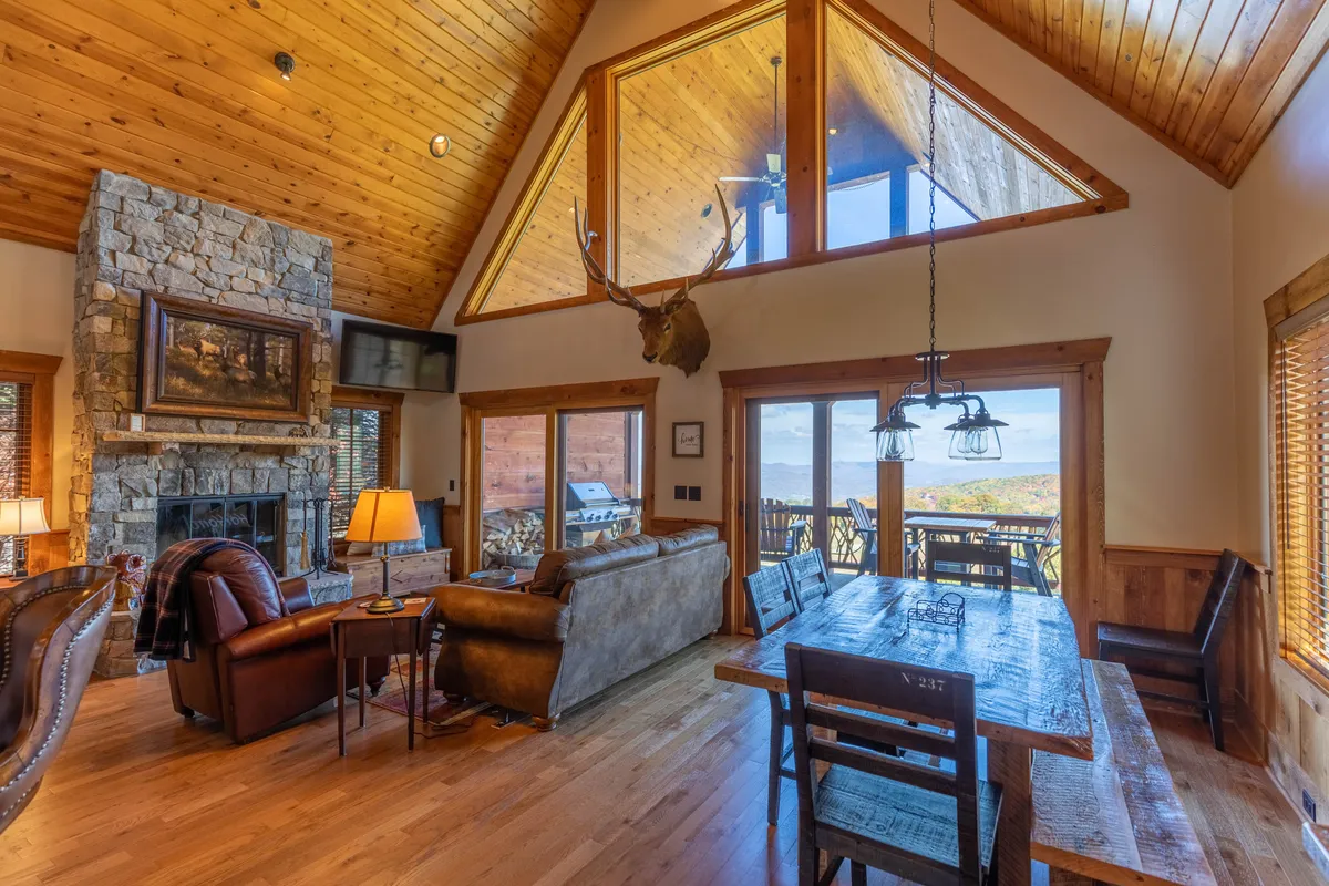 A warm, welcoming living/dining area with a substantial stone fireplace as a focal point, large picture windows offering scenic mountain views, a rustic antler chandelier, and a spacious wooden dining table set for family meals. The space radiates comfort and is well-suited for both intimate gatherings and holiday celebrations.