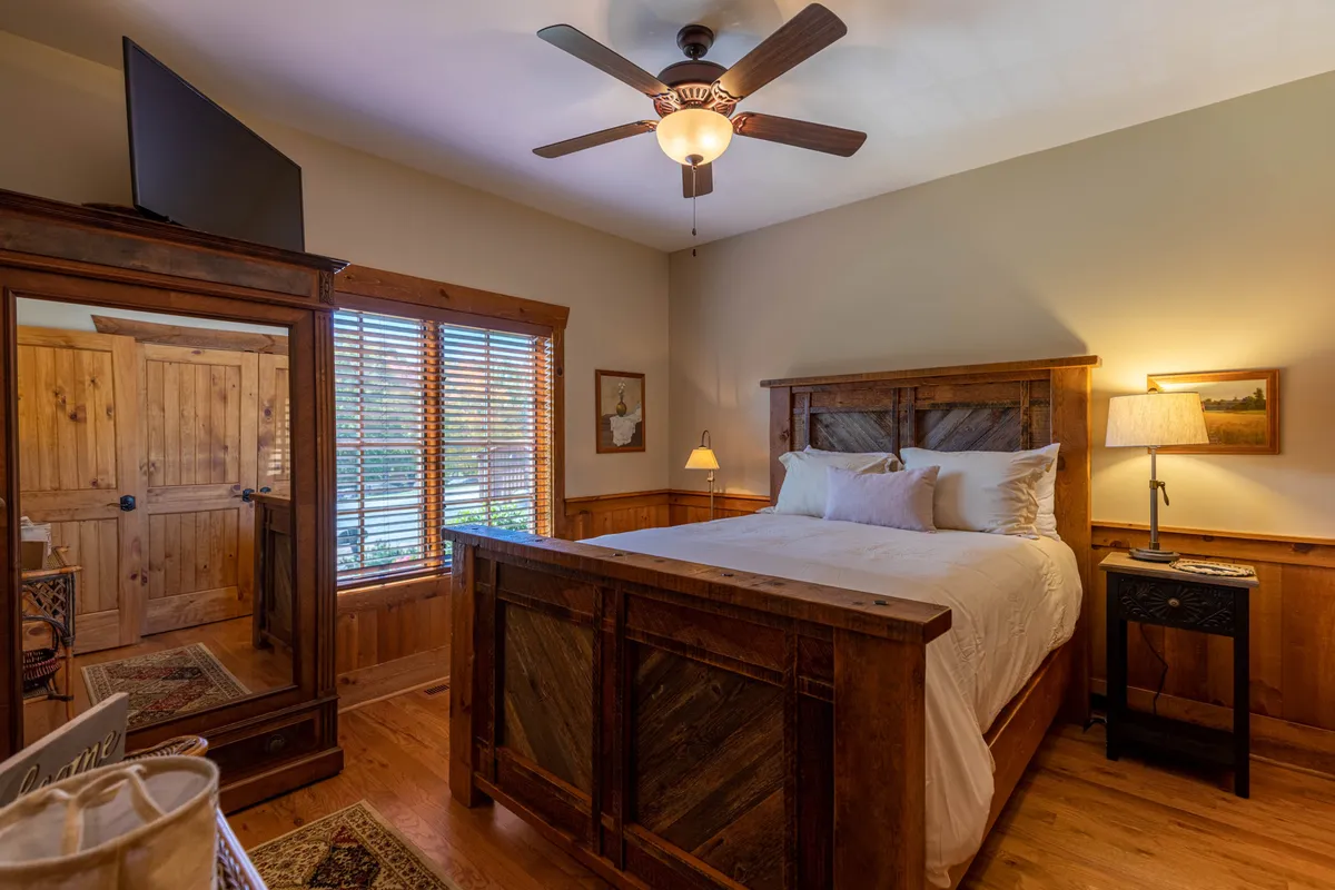 A cozy bedroom with a wooden bed frame, nightstand with lamp, wooden wardrobe, and a window with blinds. The room feels tranquil, with soft natural light filtering in, evoking a peaceful atmosphere ideal for unwinding after a busy day.