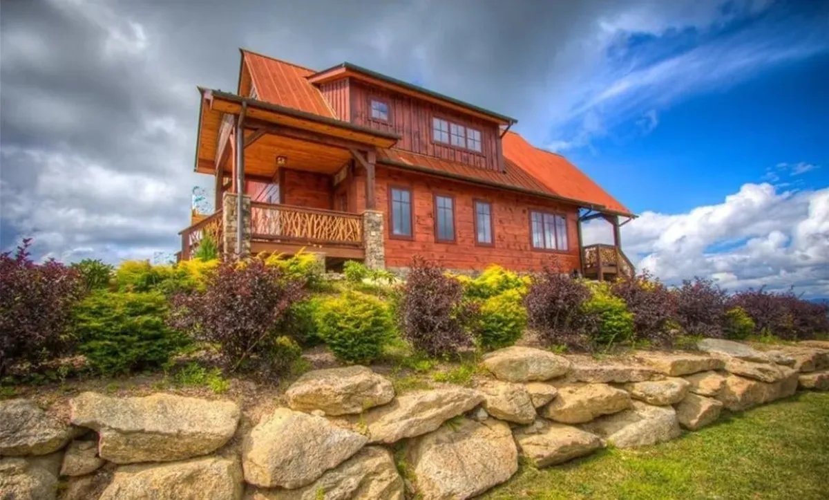 This image features a welcoming wooden house with a rock wall and lush greenery, showing a porch where families gather and relax outdoors. The setting emphasizes both the residential comfort and proximity to nature, with space for gatherings and the natural environment as a backdrop.