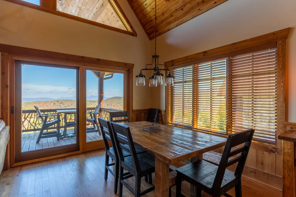 A comfortable dining area featuring a wooden table, large windows with views to the outdoors, exposed wooden beams, and warm light fixtures. The expansive windows suggest connection to natural surroundings and sunsets, making the space perfect for gatherings or quiet meals after exploring the area.
