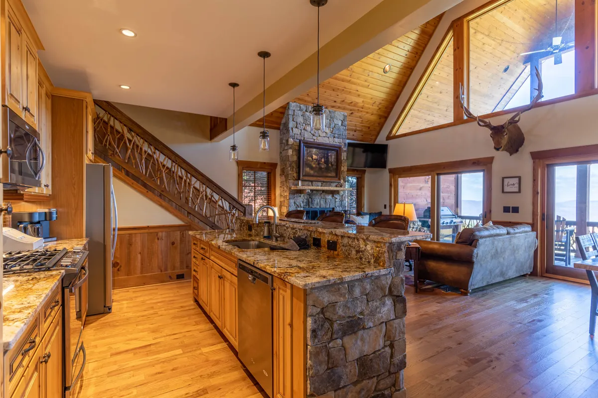 A view of the kitchen island with a stone countertop, adjacent to a cozy stone fireplace, large windows that hint at scenic mountain vistas, and a wooden staircase. The warm lighting and furnishings create an inviting space ideal for gathering after a day of exploration.