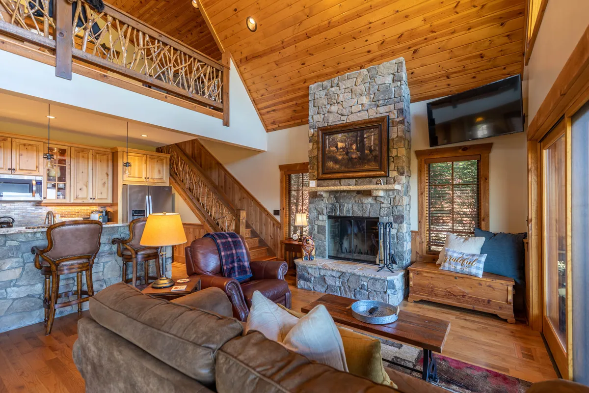 An inviting interior living area featuring a stone fireplace, exposed wooden beams, plush seating, and glimpses of the outdoors through large windows. The open plan includes a kitchen area, suggesting easy flow between indoor and outdoor spaces.
