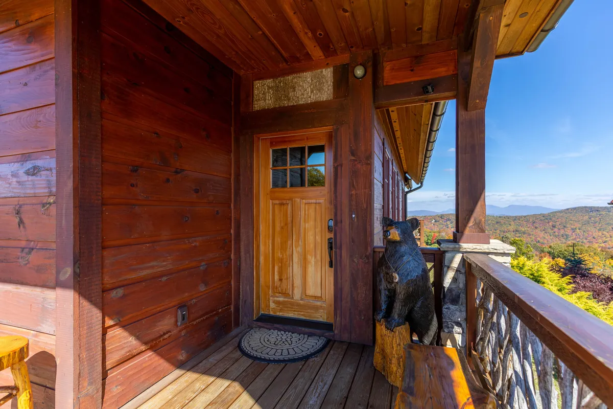 The scene centers on a rustic wooden front door framed by a friendly bear sculpture and wooden flooring, offering a sense of arrival and warm hospitality. The porch features sunrise views and hints at wildlife sightings, connecting the lodge experience to the surrounding environment.