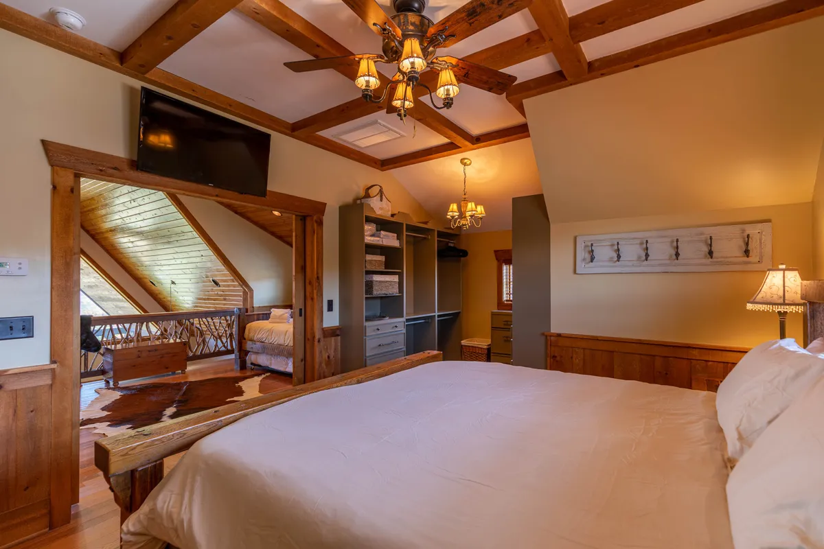 A welcoming bedroom featuring a plush bed, exposed wooden beams, a chandelier, and an open doorway leading to a loft area. The room is infused with warmth and rustic charm, with light streaming in to brighten the space.